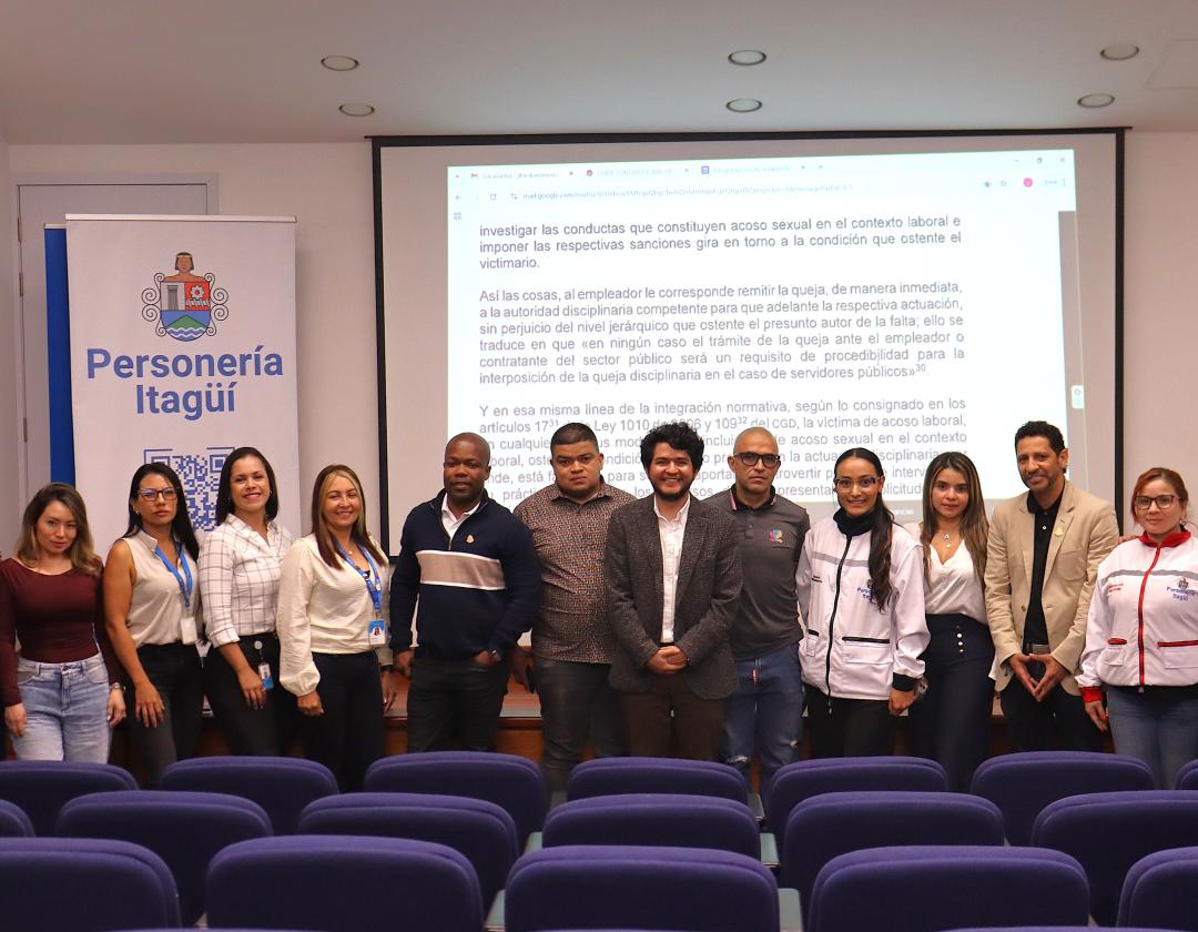 En la imagen se observa un grupo de aproximadamente 18 personas posando para la foto en lo que parece ser un auditorio o sala de conferencias. Al fondo, se ve una pantalla proyectando un texto, y a la izquierda un pendón con el logo institucional y el texto “Personería Itagüí. Las personas están alineadas al frente del auditorio, frente a varias filas de sillas azules. Se encuentran vestidos de manera formal y semiformal